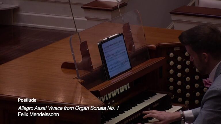 Musician playing a wooden pipe organ with a tablet displaying music on the organ bench in a formal setting