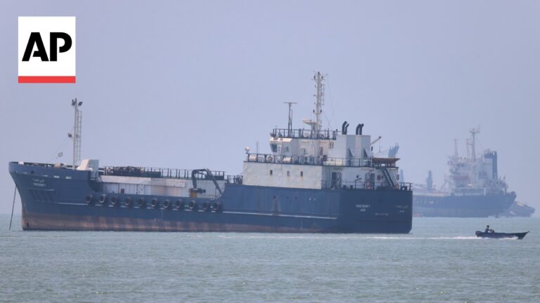 Large blue cargo ship on calm water with a small speedboat in the foreground; AP watermark in the top-left corner.