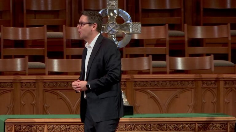 A man in a dark suit and white shirt speaking on a church stage with a large metal cross behind him.