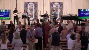 Church sanctuary with a live band on stage; banners read 'Trusting that all belong to God' and attendees mingle in the pews below.