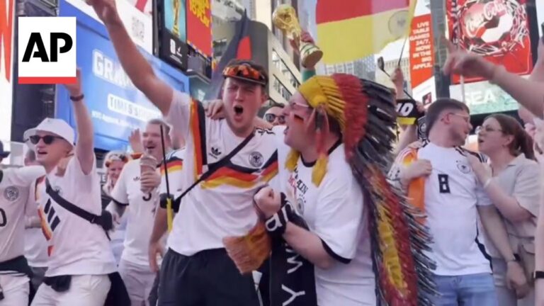 Group of Germany football fans celebrate in a busy city street, arms raised; one man wears a colorful feather headdress amid cheering teammates.