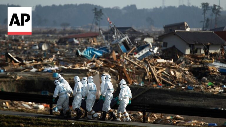 Hazmat-suited rescue workers walk along a road carrying a body on a stretcher past collapsed buildings and debris. AP logo visible in the corner.