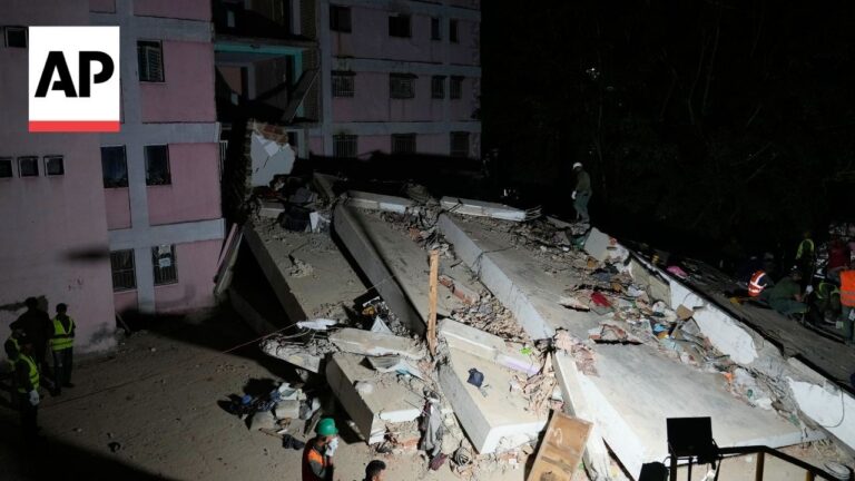 AP news photo showing a collapsed building at night with rubble and rescue workers in hard hats and vests searching the site.