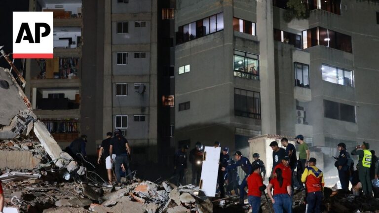 Nighttime disaster scene with a group of rescue workers sifting through rubble in front of a damaged apartment building; AP logo visible in the upper left corner.