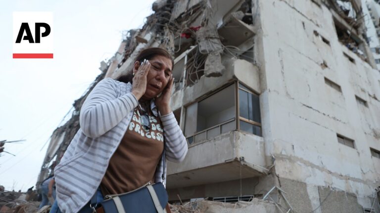 Woman in striped cardigan covers her ears amid rubble and a damaged building; AP logo visible in the corner.
