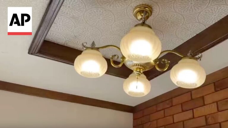 Brass chandelier with five frosted glass globe lamps hanging from a decorative ceiling near a brick wall; AP watermark in corner.