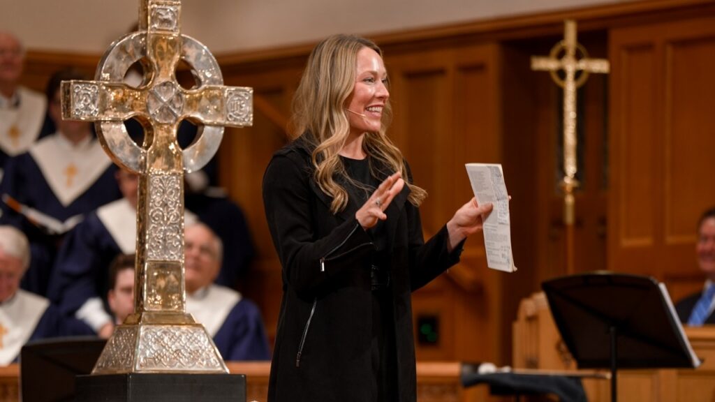 Woman in a black coat speaks at a church podium, holding a program with a silver cross and choir in the background.