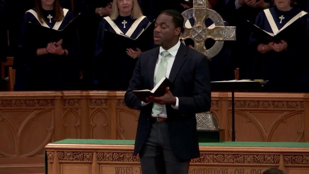 Man in a dark suit reading from a Bible at the pulpit, with a choir in blue robes behind him.