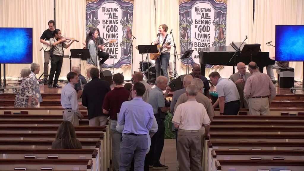 A live church band performs on stage with a guitarist, violinist, and singer while people in the pews stand and chat in the foreground.