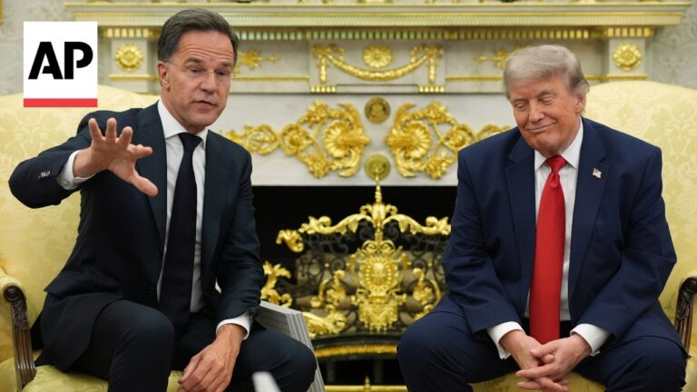 Two men in dark suits sit on gold-patterned chairs in a room with ornate gold decor; one hand raised as if explaining something, the other looking down with a slight smile; AP logo in the top left corner.