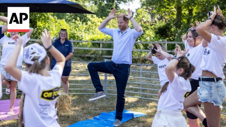 Instructor in a light blue shirt leads children in an outdoor balance exercise, arms raised, on blue mats beside a fence and trees.