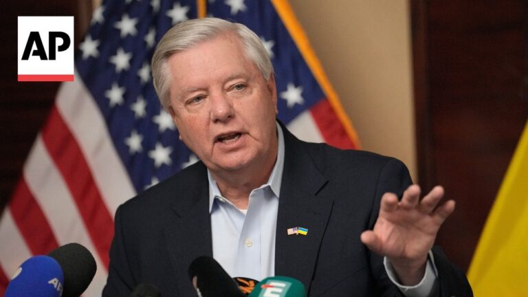 Man in a dark suit speaks at a press briefing with multiple microphones; American flags in the background and AP logo visible on the left.
