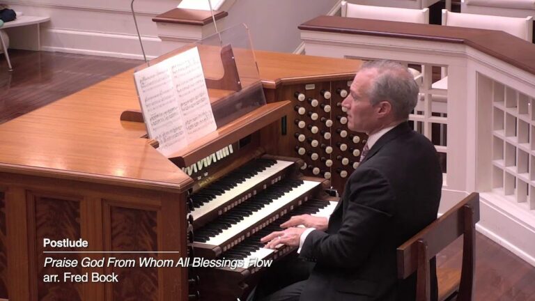 Man in a suit playing a wooden pipe organ in a church; sheet music on the stand, with on-screen caption: 'Postlude Praise God From Whom All Blessings Flow arr. Fred Bock'