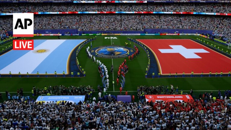 Aerial view of a stadium field with large Argentina (left) and Switzerland (right) banners on the pitch and teams lined up around a central emblem, crowd in stands.