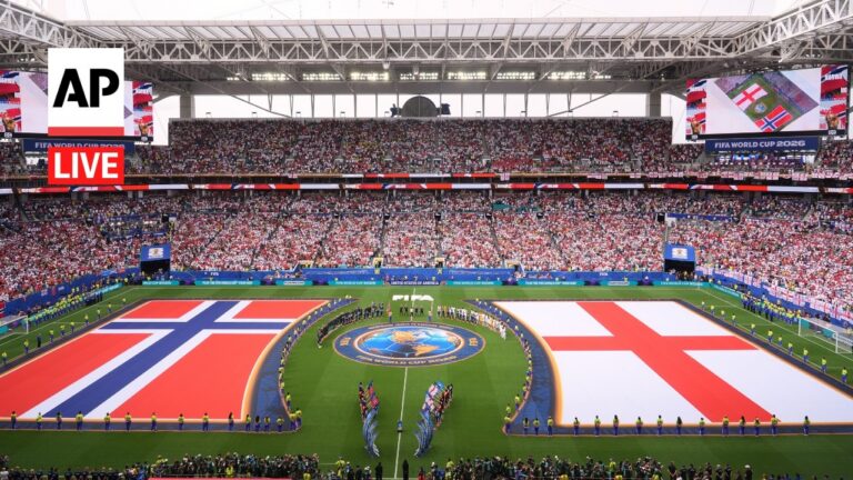 Stadium filled with spectators as two large national flags (Norway left, England right) cover the field for a World Cup 2026 ceremony, center emblem visible, security lining the pitch edges.