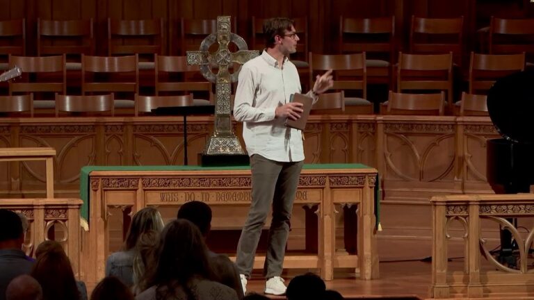 Man in a white shirt speaks to an audience on a stage in a church, holding a tablet, with a decorative cross behind him.