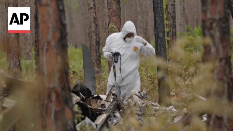 Person in a white hazmat suit in a forest, kneeling among wreckage and debris, holding a camera with a strap, AP logo in the upper left corner.
