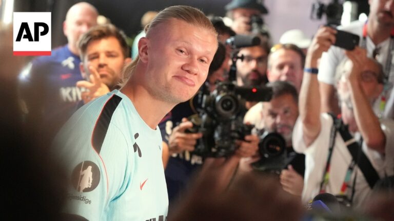 Smiling athlete in a light blue jersey surrounded by reporters and photographers at a press event.