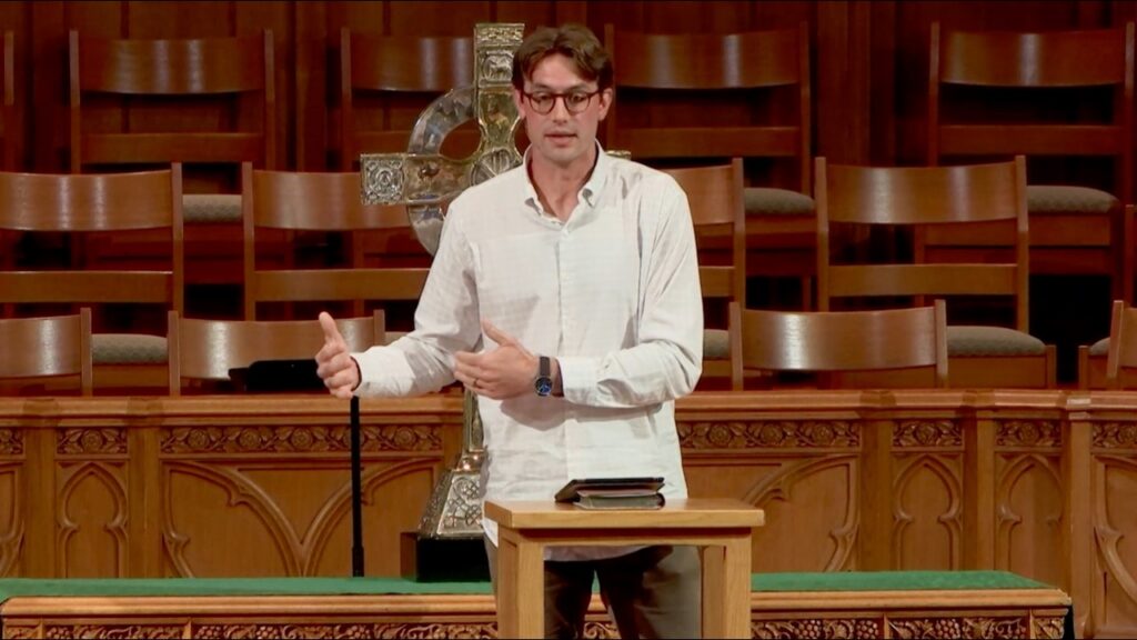 1785314478_maxresdefaultjpg - New Social Gospel Man in a white shirt speaks from a wooden pulpit in a church, with a cross and empty pews behind him.