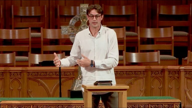 Man in a white shirt speaks from a wooden pulpit in a church, with a cross and empty pews behind him.