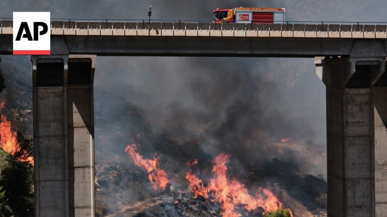 Fire truck on a highway bridge above a wildfire, with orange flames and dense smoke below.