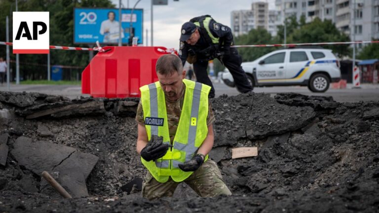 Police officer in a bright vest stands in a rubble-filled trench, inspecting debris; a second officer and a police car are in the background behind barriers.