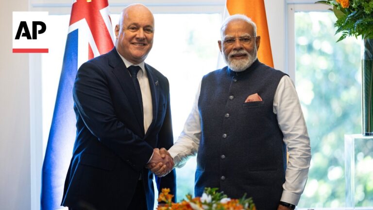 Two men in suits shake hands in a bright room, flags in the background and an AP logo in the corner.
