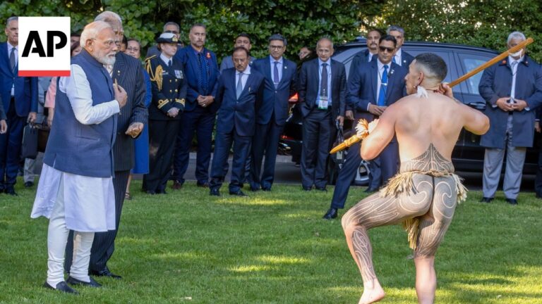 Prime Minister Narendra Modi and other officials watch a shirtless, intricately tattooed dancer performing with a staff on a grassy lawn.