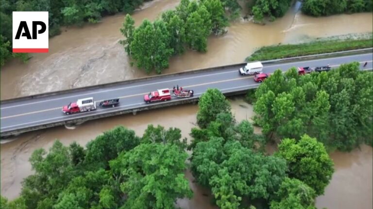 Aerial view of a flooded highway with water surrounding the bridge and vehicles, including a red truck and emergency responders on the roadway (AP logo top-left).