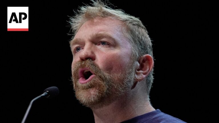 Close-up of a bearded man speaking into a microphone on stage; AP logo in the top-left corner.