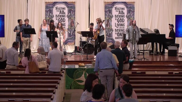 Worship band performs on a church stage with singers, guitarist, drummer, and pianist; audience mingle in front.