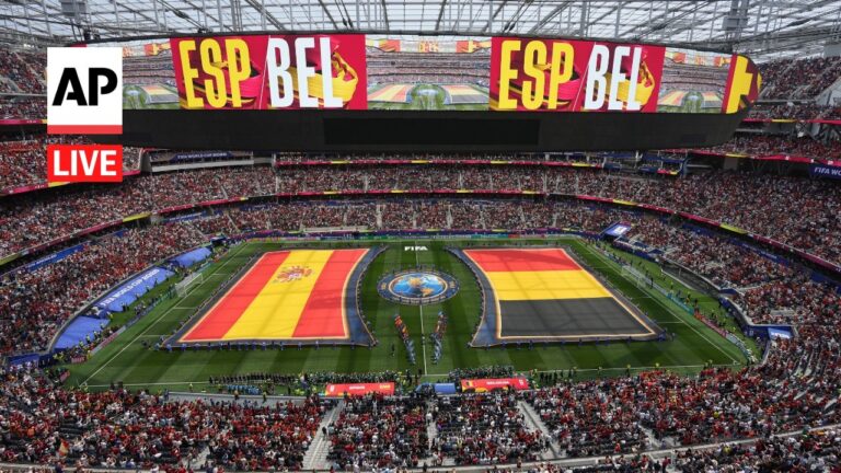 Packed stadium with a live broadcast overlay; two oversized Spain flags span the pitch amid a cheering crowd and a central emblem on the field.