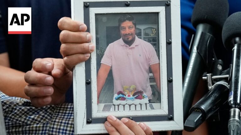 Framed photo of a man in a pink polo with a birthday cake showing 52, held at a press conference beside microphones.