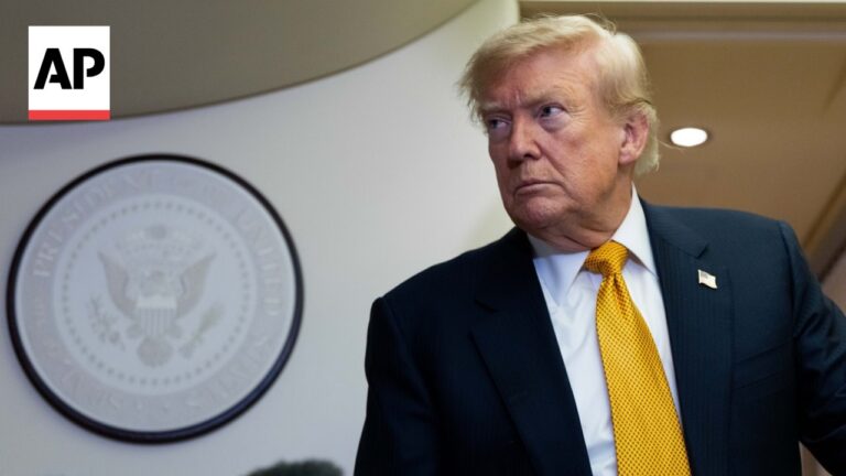 Man in a dark suit with a yellow tie stands beside a circular seal on a wall, AP logo in the upper left corner.