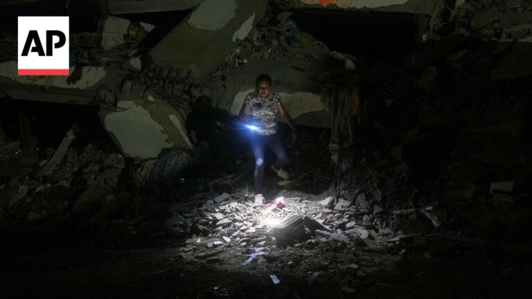 Person in a patterned shirt searches through rubble at night, flashlight lighting debris after a collapse; AP logo in corner.