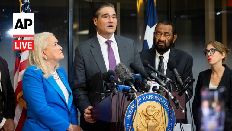 Man in a gray suit speaks at a press conference from a podium with multiple microphones, flanked by a blonde woman in a blue blazer and two colleagues.