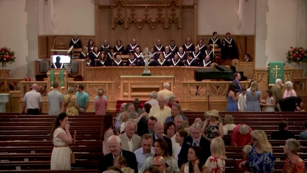 Choral group in black robes with white trim sings on an elevated platform in a church, with congregants mingling in the pews below.