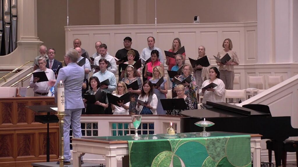 A choir stands on a church balcony led by a conductor in a light-blue suit, with a grand piano nearby and a candle on the altar in front of a green-cloth table.