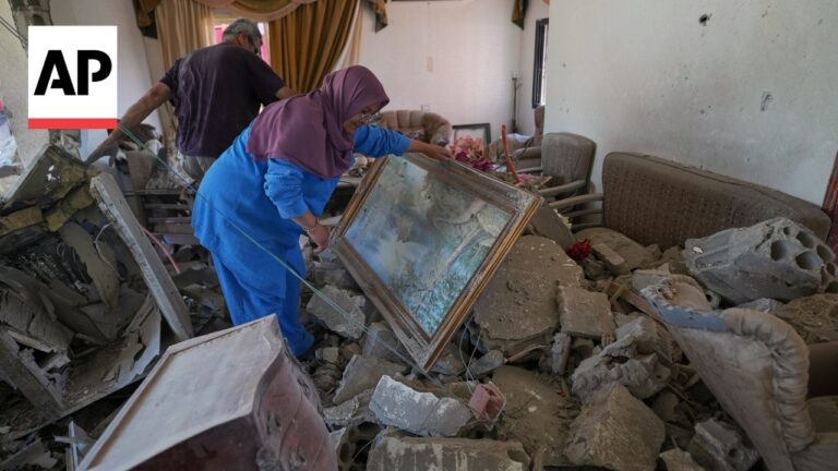 AP News logo in upper-left corner; a woman in a purple hijab and blue outfit lifts a framed painting from rubble inside a damaged room, with a man in the background.