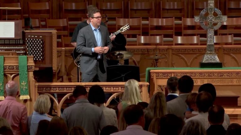 Man in a gray suit plays a guitar at the church podium while the congregation faces the front, with ornate woodwork and a cross on the altar behind.