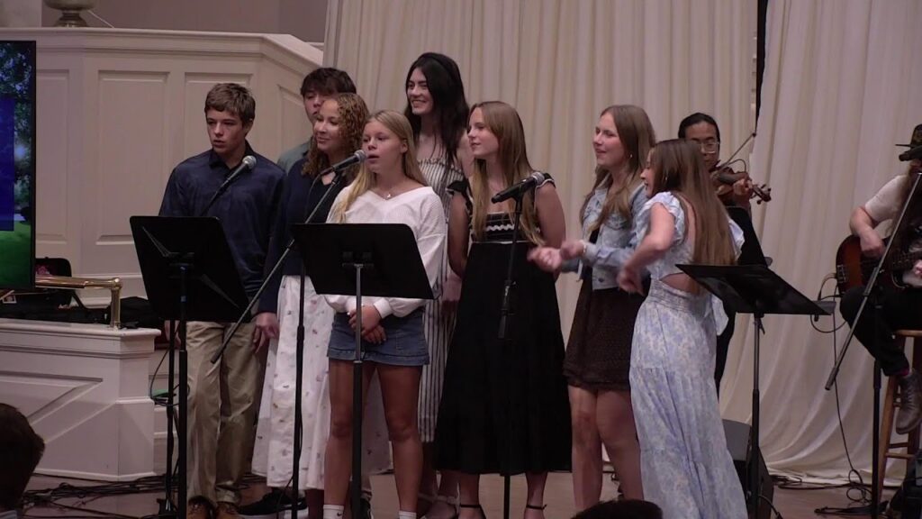 A group of young singers performing on a stage with microphones, dressed casually and in dresses, with musicians in the background.