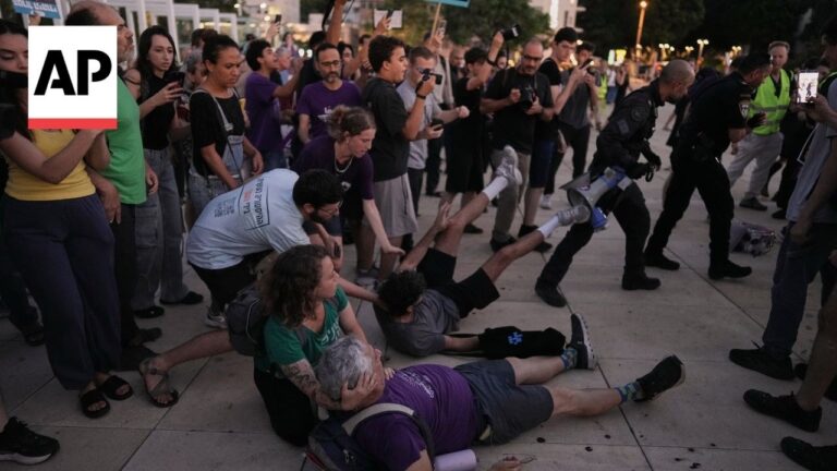 Crowd of bystanders assists injured protesters on a sidewalk while police monitor the scene at dusk, many filming with phones.