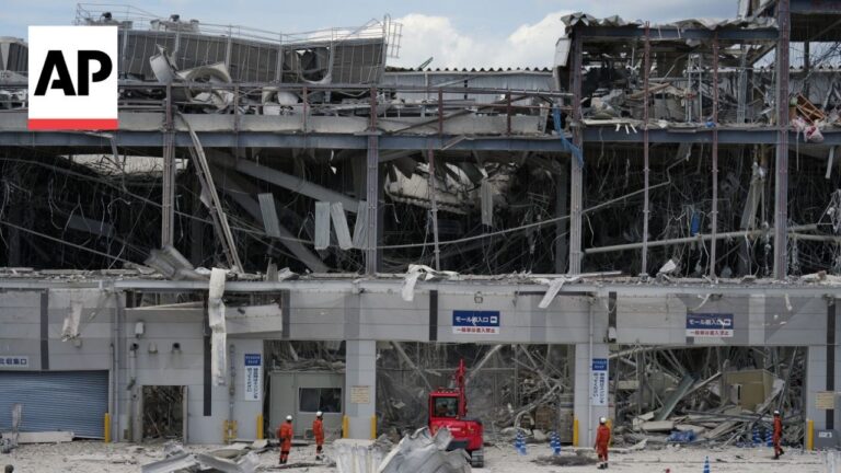 AP logo beside a collapsed, debris-strewn building with exposed steel and rubble, as orange-suited workers survey the scene.