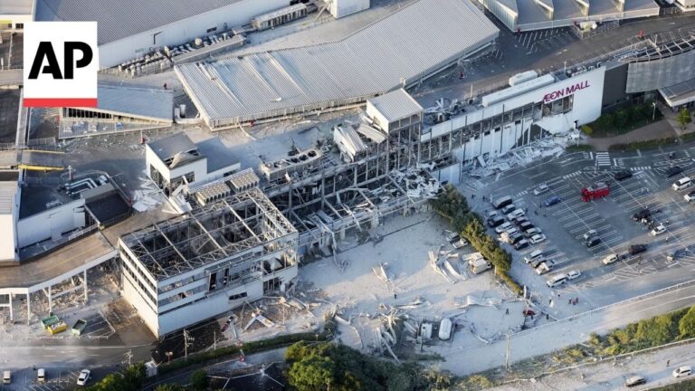 Aerial view of a damaged AEON Mall with collapsed roofs, exposed framework, and scattered debris in a parking lot, AP logo in the corner.