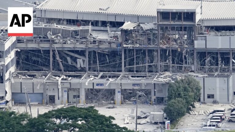 AP logo in top-left corner over a collapsed, debris-filled building facade and shattered windows; foreground shows trees and a row of parked cars.