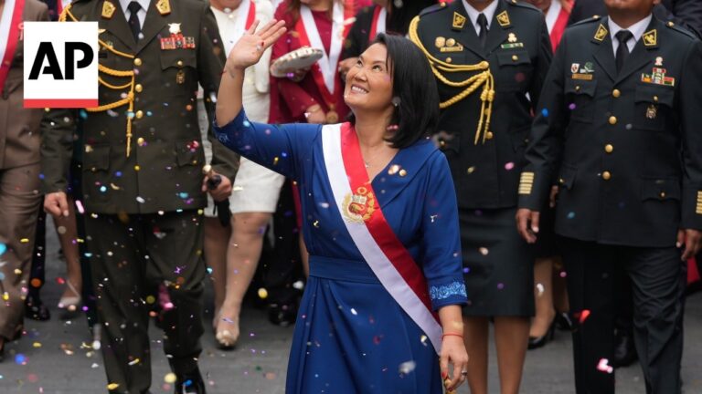 Woman in a blue dress with a red-and-white sash waves and smiles amid a confetti shower, with uniformed soldiers behind. (AP logo by left edge)