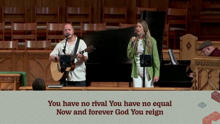1786897199_maxresdefaultjpg - New Social Gospel Two worship musicians perform on a church stage: a man with an acoustic guitar and a woman singing into a microphone, with a pianist in the background and lyric banner in front of the pews.