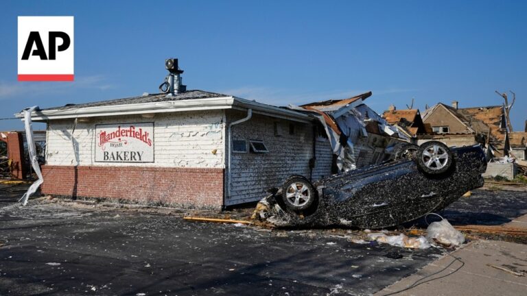 AP News logo on the left; a toppled car lies upside down in front of a damaged bakery building with a sign reading 'Manderfields Home Bakery' under a blue sky.
