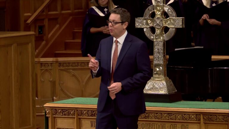 A man in a navy suit giving a speech at a wooden pulpit, with a large ornate silver cross behind him in a church setting.
