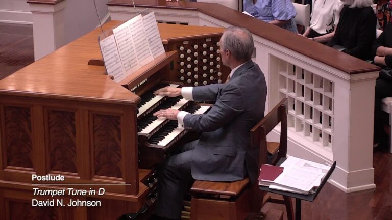 Organist in a gray suit playing a wooden pipe organ; on-screen caption reads 'Postlude: Trumpet Tune in D — David N. Johnson'.